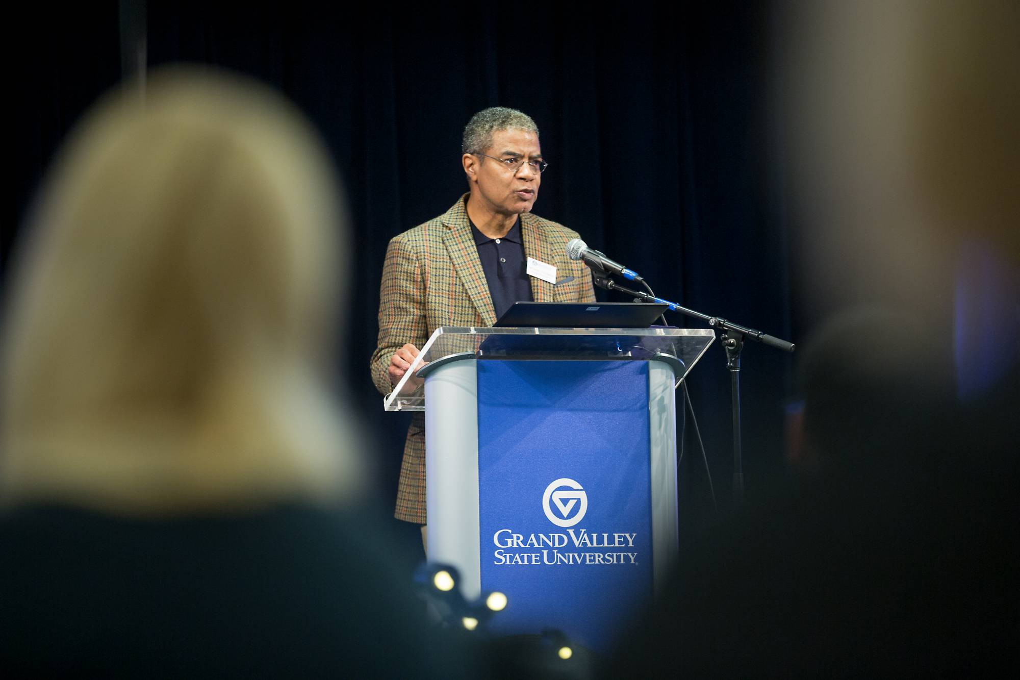 A man standing on a stage and talking into a microphone. There is a podium in front of him.